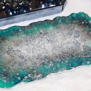 Puede incluir: Una bandeja de servir de resina turquesa y plata con forma libre y acabado brillante. La bandeja tiene dos pequeños pomos de cristal en el lateral.