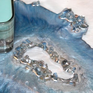 May include: A decorative resin tray with a coastal theme. The tray features shades of blue and white, with embedded glass pieces and a central white element. A glass candle holder with a seashell is visible.