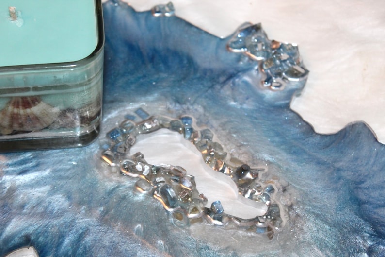 May include: A decorative resin tray with a coastal theme. The tray features shades of blue and white, with embedded glass pieces and a central white element. A glass candle holder with a seashell is visible.