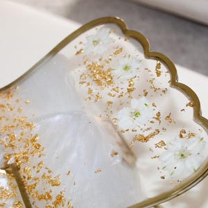 May include: A decorative tray with a scalloped edge, featuring a white and gold design. The tray is filled with white flowers and gold flakes, encased in a clear resin. The edge of the tray is trimmed with gold.