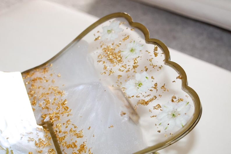 May include: A decorative tray with a scalloped edge, featuring a white and gold design. The tray is filled with white flowers and gold flakes, encased in a clear resin. The edge of the tray is trimmed with gold.