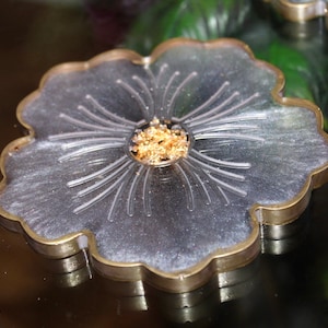 May include: A silver and gold resin coaster with a flower design. The coaster has a raised center with gold flakes.