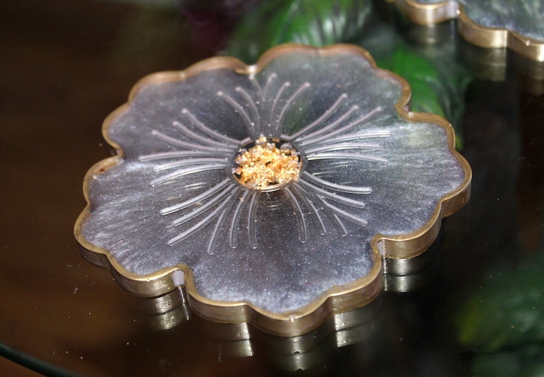 May include: A silver and gold resin coaster with a flower design. The coaster has a raised center with gold flakes.