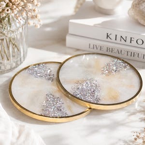 May include: Two round coasters with a gold rim. The coasters have a white and gold background with silver glitter accents. The coasters are on a white surface with a vase of flowers and books in the background.