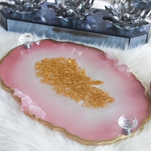 May include: A pink and gold resin tray with crystal handles. The tray has a gold edge and a gold glitter design in the centre. Decorative items, including baubles and candle holders, are in the background.
