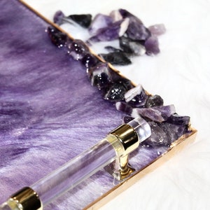 May include: A purple resin tray with a gold rim and a clear acrylic handle. The tray is decorated with amethyst crystals.