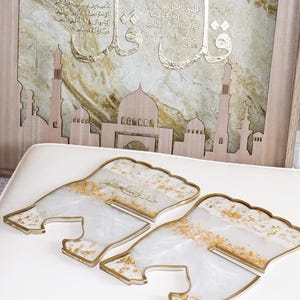 May include: Decorative Islamic art piece featuring a wooden mosque silhouette and a framed artwork with Arabic calligraphy in gold. Four clear, resin-like coasters with gold accents are in the foreground.
