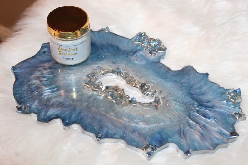 May include: A decorative resin tray with a geode-like design in shades of blue and silver. The tray has a unique, organic shape and is adorned with crystal accents. A jar with a gold lid and the text "Aqua Surf Surf aqua" is on the tray.