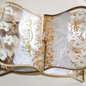 May include: A decorative, open book-shaped tray with a gold-colored frame. The clear resin is embedded with white flowers and gold flakes. The tray has Arabic script in gold lettering.