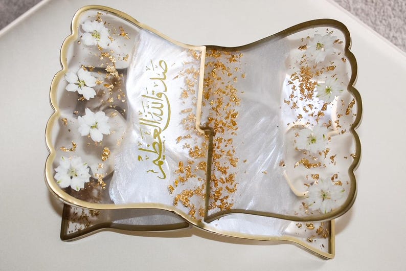May include: A decorative, open book-shaped tray with a gold-colored frame. The clear resin is embedded with white flowers and gold flakes. The tray has Arabic script in gold lettering.