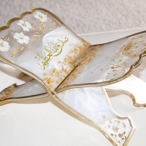 May include: An ornate, open book stand with a scalloped edge. The stand is white with gold accents, including gold leaf and a gold border. White flowers are embedded in the surface. Arabic script is visible on the surface.