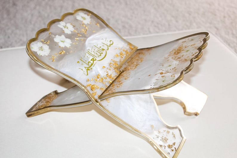 May include: An ornate, open book stand with a scalloped edge. The stand is white with gold accents, including gold leaf and a gold border. White flowers are embedded in the surface. Arabic script is visible on the surface.