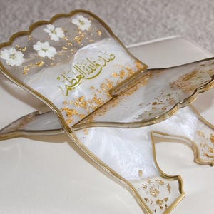 May include: A decorative book stand made of resin, featuring a white and gold color scheme. The stand has a scalloped edge and is adorned with white flowers and gold leaf accents. Arabic script is visible on the surface.