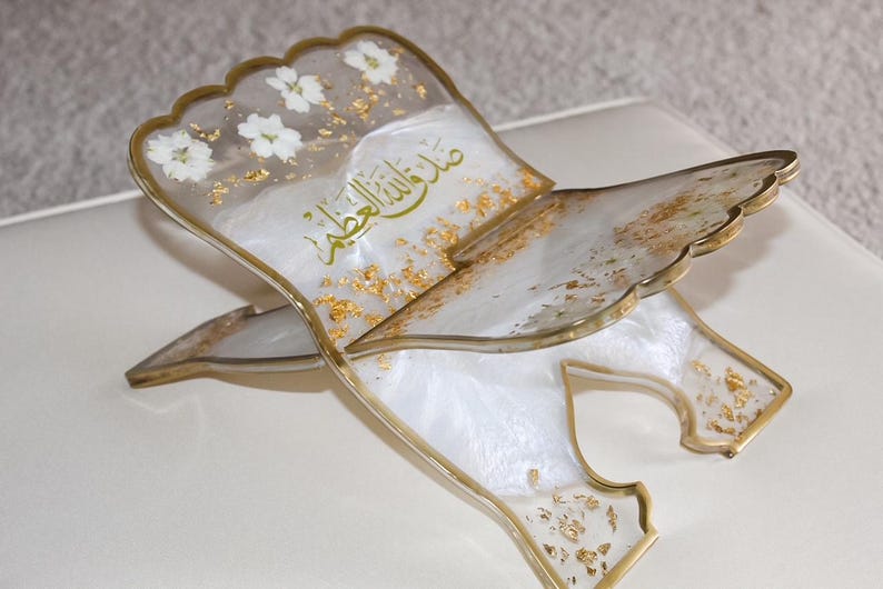 May include: A decorative book stand made of resin, featuring a white and gold color scheme. The stand has a scalloped edge and is adorned with white flowers and gold leaf accents. Arabic script is visible on the surface.