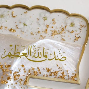 May include: Decorative resin tray with a scalloped edge, featuring white flowers, gold leaf accents, and Arabic calligraphy. The tray has a pearlescent white base and a gold-colored trim.