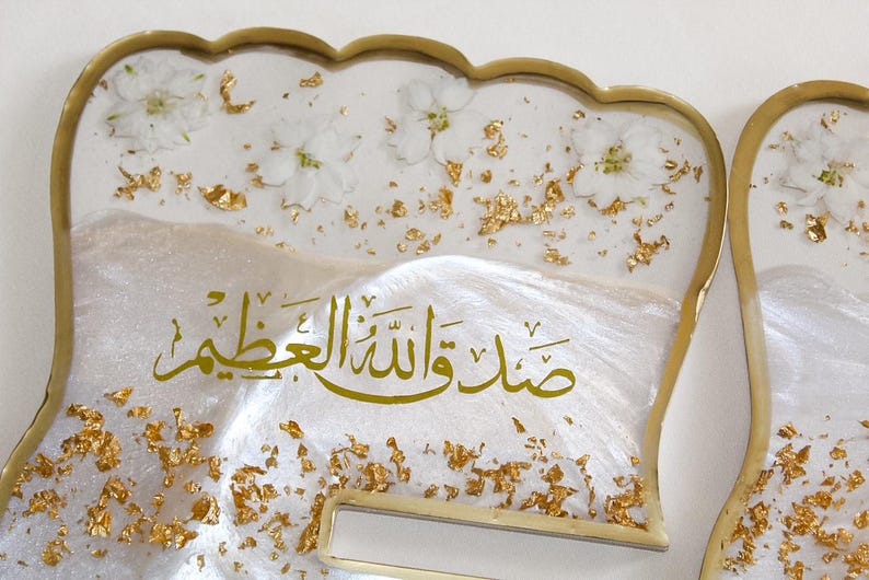 May include: Decorative resin tray with a scalloped edge, featuring white flowers, gold leaf accents, and Arabic calligraphy. The tray has a pearlescent white base and a gold-colored trim.