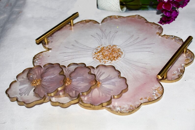 May include: A set of four pink and gold resin coasters with a flower design. The coasters are arranged in a stack, with the largest coaster on the bottom and the smallest coaster on top. The coasters have a gold rim and a flower design in the center. The coasters are perfect for protecting your furniture from scratches and spills.