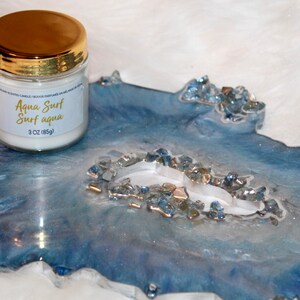 May include: A scented candle in a glass jar with a gold lid, labeled "Aqua Surf Surf aqua" and 3 oz (85g). It sits on a blue and white resin tray with a geode-like design, accented with blue and silver crystals.