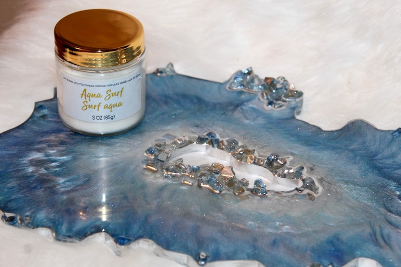 May include: A scented candle in a glass jar with a gold lid, labeled "Aqua Surf Surf aqua" and 3 oz (85g). It sits on a blue and white resin tray with a geode-like design, accented with blue and silver crystals.