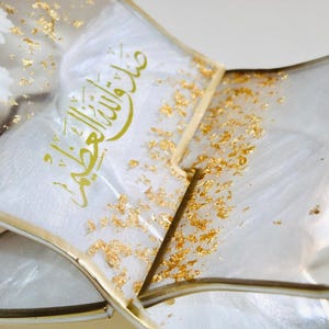 May include: Close-up of a decorative, clear resin tray with gold accents and gold leaf flakes. White flowers are embedded in the resin. The tray contains a white fabric with gold Arabic calligraphy.