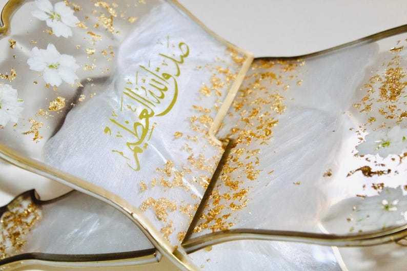 May include: Close-up of a decorative, clear resin tray with gold accents and gold leaf flakes. White flowers are embedded in the resin. The tray contains a white fabric with gold Arabic calligraphy.