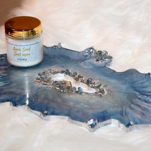 May include: A decorative resin tray in shades of blue and silver, with a geode-like design. A small jar with a gold lid sits on the tray, labeled "Aqua Surf" and "3 oz (85g)". The tray has a unique, organic shape.