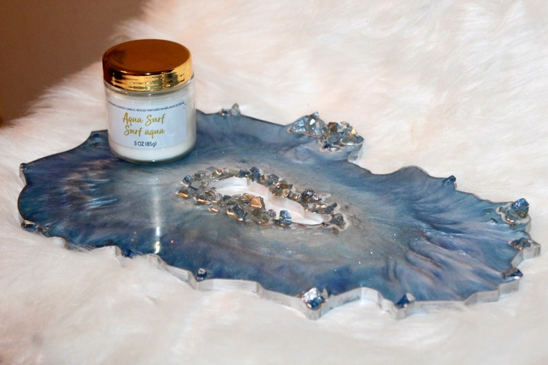 May include: A decorative resin tray in shades of blue and silver, with a geode-like design. A small jar with a gold lid sits on the tray, labeled "Aqua Surf" and "3 oz (85g)". The tray has a unique, organic shape.