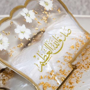 May include: A decorative book stand with a clear resin design. The stand features white flowers, gold leaf accents, and gold lettering. The stand has a scalloped edge and a gold-colored frame.