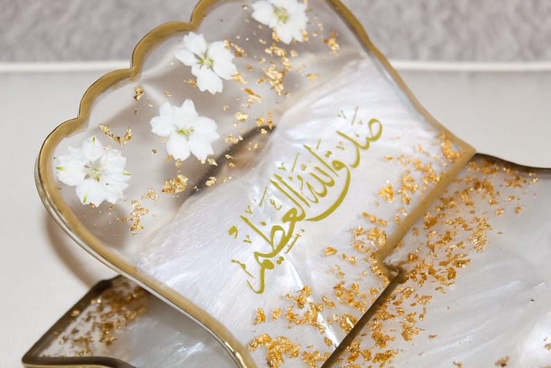 May include: A decorative book stand with a clear resin design. The stand features white flowers, gold leaf accents, and gold lettering. The stand has a scalloped edge and a gold-colored frame.