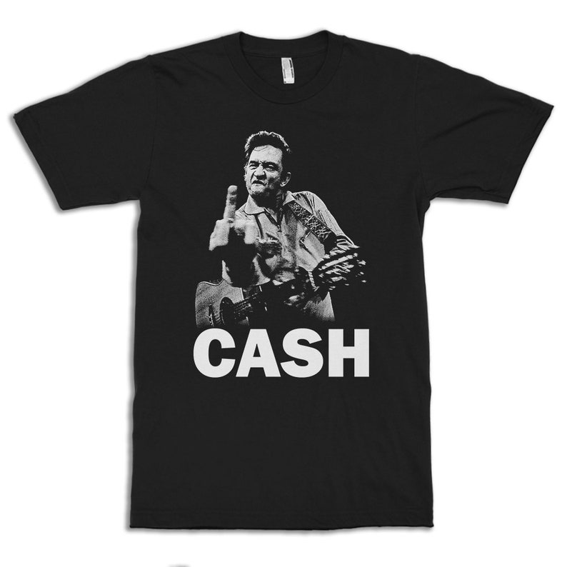Puede incluir: Camiseta negra con una imagen blanca de Johnny Cash haciendo el dedo medio mientras toca la guitarra. El texto "CASH" est&aacute; impreso en blanco debajo de la imagen.