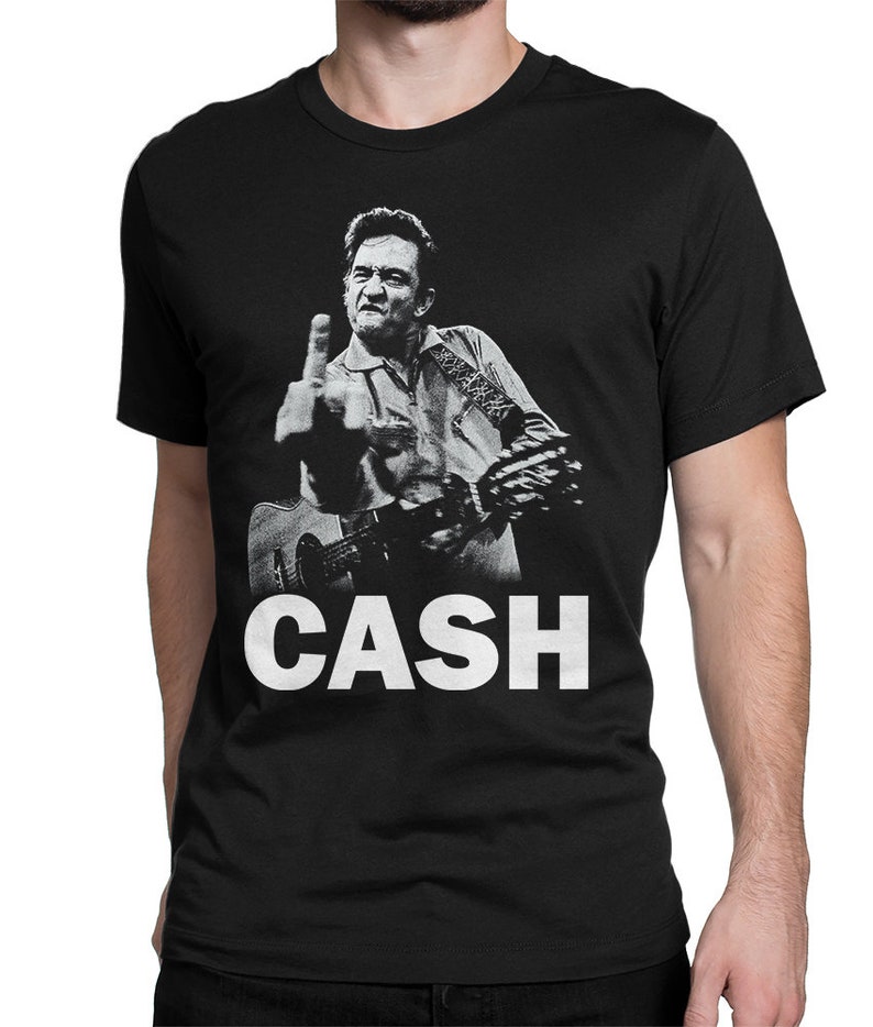 Puede incluir: Camiseta negra con una imagen en blanco y negro de Johnny Cash haciendo el dedo medio mientras toca la guitarra. El texto "CASH" est&aacute; impreso en blanco debajo de la imagen.