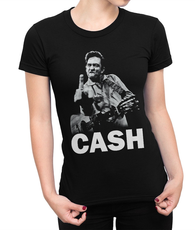 Puede incluir: Camiseta negra con una foto en blanco y negro de Johnny Cash haciendo el dedo medio mientras toca la guitarra. El texto "CASH" est&aacute; impreso en blanco debajo de la foto.