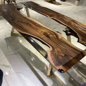 Puede incluir: Una gran mesa de comedor de madera con una incrustación de resina epoxi transparente. La mesa tiene un acabado de madera natural y una base de metal dorado.