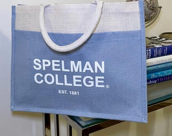 May include: A blue and white tote bag with the words "SPELMAN COLLEGE" and "EST. 1881" printed in white. The bag has white handles and is made of a textured material. The bag is sitting on a glass table.