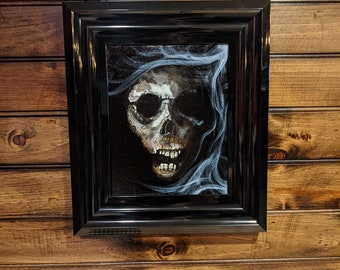 May include: A framed painting of a skull with a dark background and white wisps. The skull is depicted with a gaping mouth and visible teeth. The frame is black and glossy, mounted on a wooden wall.
