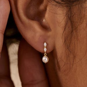 May include: A gold drop earring featuring two small marquise-cut clear stones stacked vertically, connected to a single round white pearl dangling at the base. The earring is secured to the earlobe with a thin gold post and backing mechanism.