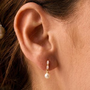 May include: A gold drop earring featuring a small, round white pearl suspended from a thin gold bar accented with two tiny, clear, marquise-cut stones. The earring is secured to the earlobe with a simple gold post and backing mechanism.