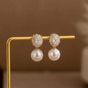 May include: A pair of gold-tone earrings featuring a central oval-cut clear gemstone surrounded by a halo of small baguette-cut stones, with a smooth white spherical pearl drop suspended from the base of each earring setting.