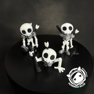 Könnte beinhalten: Drei weiße und graue Skelettfiguren mit schwarzen Waffen und Rüstungen werden auf einer schwarzen Oberfläche ausgestellt. Ein Skelett sitzt, ein anderes steht und das dritte liegt. Ein schwarzer Ninja-Stern ist ebenfalls vorhanden.