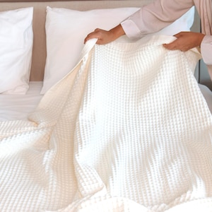 May include: A cream-coloured waffle-weave blanket being arranged on a bed with white pillows. The blanket has a textured, grid-like pattern. The bed has a neutral-toned headboard.
