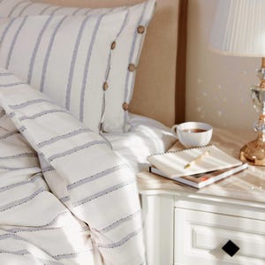 Può includere: Set di biancheria da letto bianco con motivo a righe blu. Il cuscino presenta bottoni in legno. Un comodino bianco ospita una lampada, una tazza di tè e un taccuino con una penna. La lampada ha una base di cristallo e un paralume bianco.