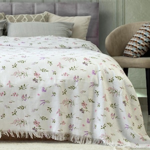 Könnte beinhalten: Weißes Bettlaken mit floralem Muster aus rosa und lila Blumen und grünen Blättern. Das Bettlaken hat einen Fransenrand und ist über ein Bett drapiert. Das Bettlaken besteht aus einem strukturierten Stoff.