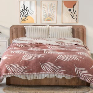 Puede incluir: Una cama con cabecero de madera y una manta marrón rojiza con un estampado de hojas blancas. Dos almohadas a rayas y tres obras de arte abstractas enmarcadas cuelgan sobre la cama. Una pequeña mesita de noche blanca y una alfombra redonda de yute también son visibles.