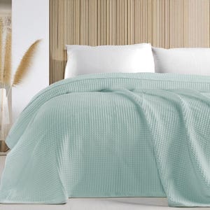 May include: A light blue waffle-weave blanket draped over a bed with white pillows. The blanket has a textured surface and is made of a soft material. The background features a wooden panel wall.