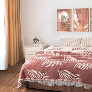 Puede incluir: Una escena de dormitorio con una cama cubierta por una manta marrón rojiza adornada con patrones de hojas blancas. La cama está hecha con almohadas a rayas y una sábana blanca. Dos obras de arte enmarcadas cuelgan sobre la cama. También se ve una mesita de noche blanca y cortinas color melocotón.