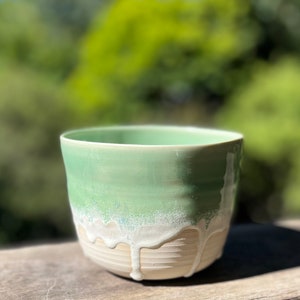 May include: A ceramic cup with a light green glaze and a white drip glaze. The cup is sitting on a wooden surface.