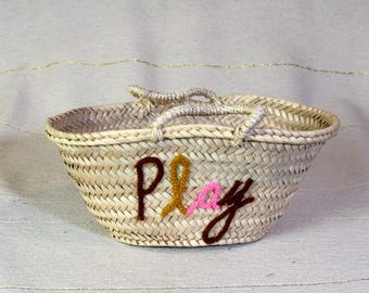 Könnte beinhalten: Eine geflochtene Strohtasche mit einer Stickerei aus braunem, rosa und gelbem Garn, die "Play" lautet.