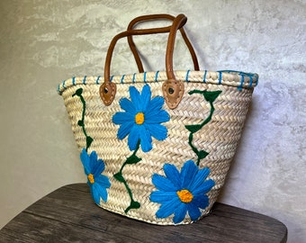 May include: A woven straw tote bag with blue and green embroidered flowers. The bag has leather handles and a blue and white trim.