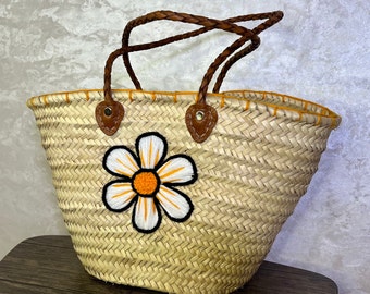May include: A straw tote bag with a brown leather handle and a white daisy with an orange center embroidered on the front.