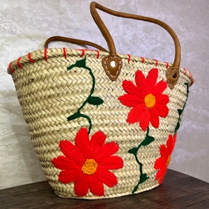 May include: A woven straw tote bag with leather handles and embroidered red flowers. The bag has a natural straw color with red and green embroidery.